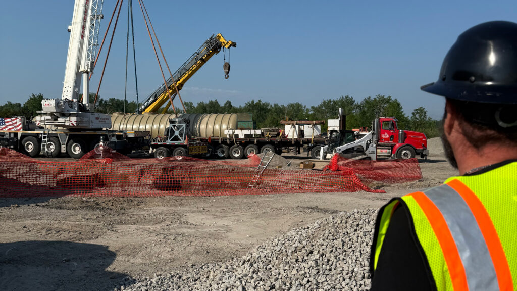 Worker in vest and hardhat looking over a tank being lowered by a crane.