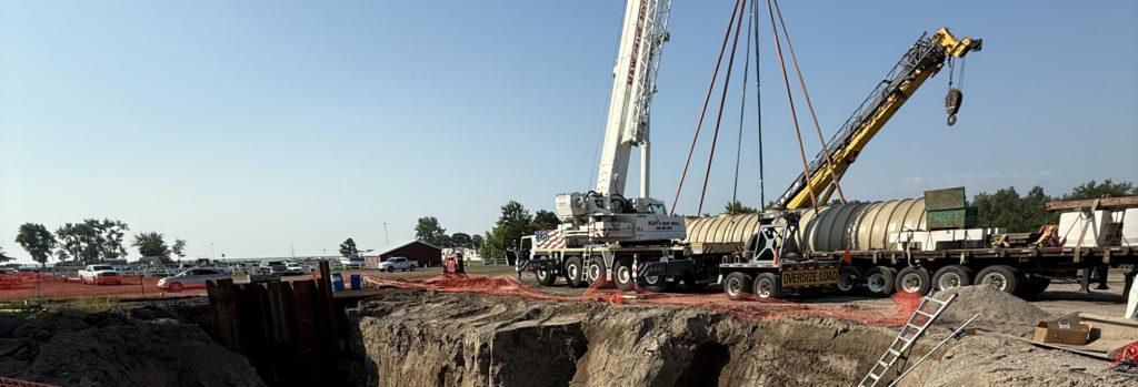 Site photo with crane and large tank behind a large hole in the ground.